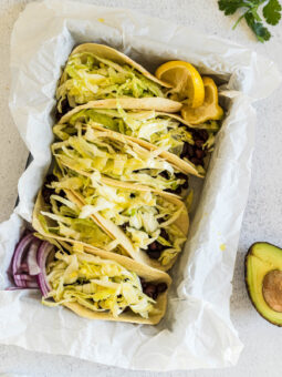 A baking dish holding 6 Vegan Black Bean Tacos With Ginger Sauce. Red onion and lemon slices are wedged next to the tacos. An avocado and cilantro are next to the tray.