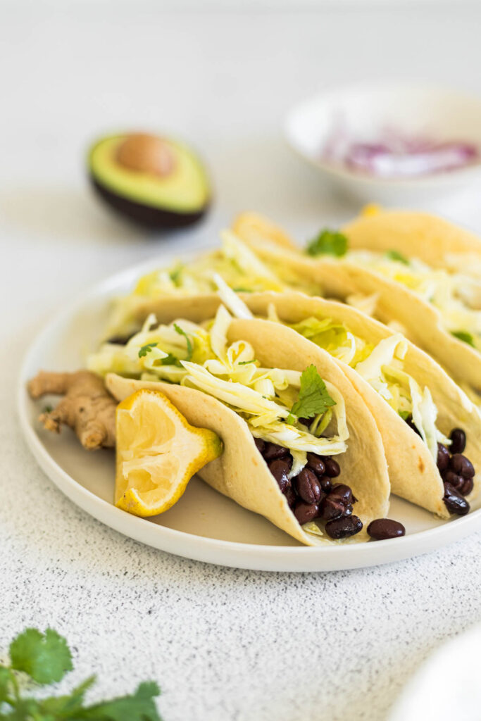 A plate of vegan black bean tacos being held up by a piece of ginger and a lemon wedge. An avocado and bowl of red onion slices sit in the background.