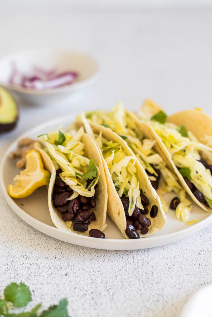 A plate of Vegan Bean Tacos. A lemon slice and ginger sit next to it. A bowl of red onion slices sit in the background, along with an avocado and cilantro.