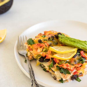 A big slice of Vegetarian Mexican Lasagna is on a plate with lemon slices and avocado on top. A fork sits next to it. Some lemon and avocado, as well as another plate of lasagna sit in the background.