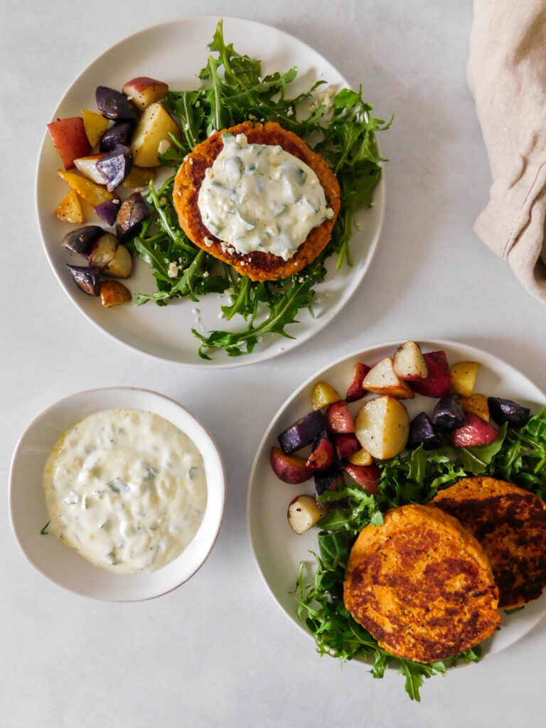 An overhead view of Sun-Dried Tomato Chickpea Patties on two plates with arugula and roasted potatoes. A bowl of yogurt dipping sauce sits on the side. 