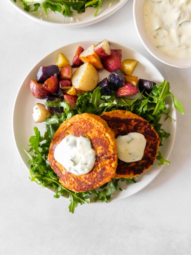 An overhead view of two chickpea patties on a plate with arugula and roasted potatoes. A bowl of yogurt dipping sauce sits on the side. 