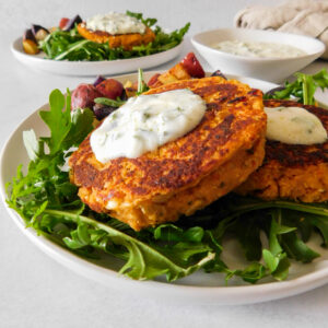 A Sun-Dried Tomato Chickpea Patty sits on top of another. They both are topped with a yogurt sauce and are sitting on top of a bed of arugula. Another plate with patties sits in the background, along with a small bowl with yogurt sauce.