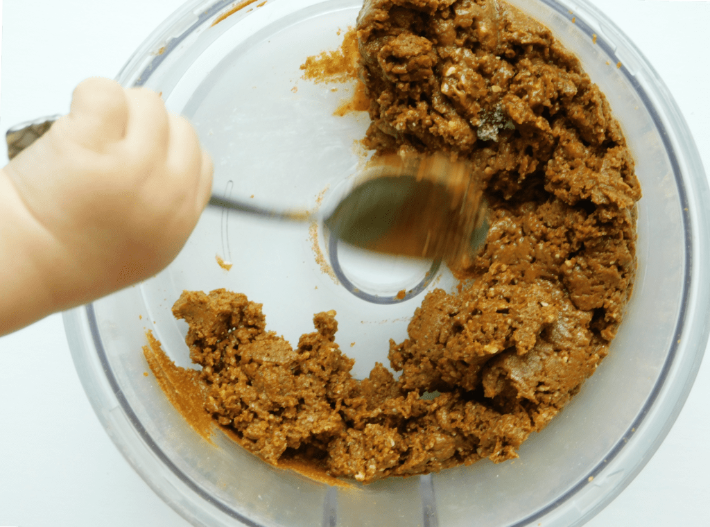A toddler hand taking a spoon to scoop out the almond butter filling from a food processor.