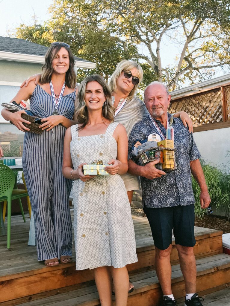 Kaileigh, a white woman, is standing on a raised back porch with a medal around her neck and is holding the first place prizes she won. Two more white women, who won second prize, stand next to Kaileigh. An older white man who won third place stands next to them holding his prizes.