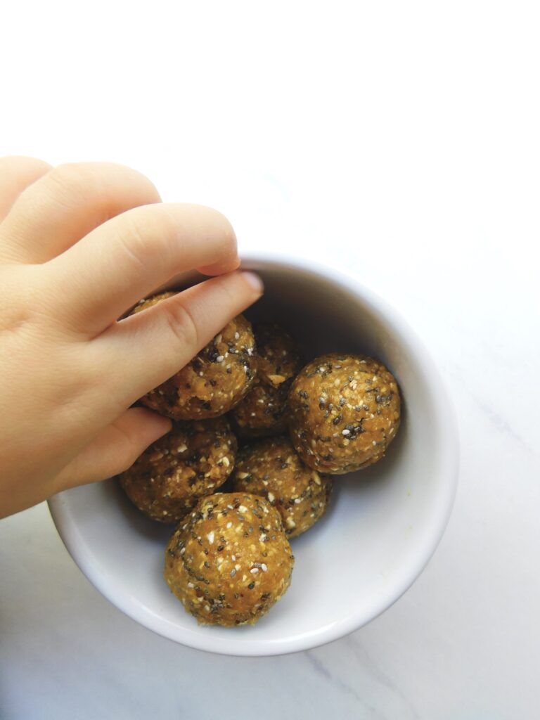 A toddler hand is reaching for an apricot energy ball in a small bowl filled with them.