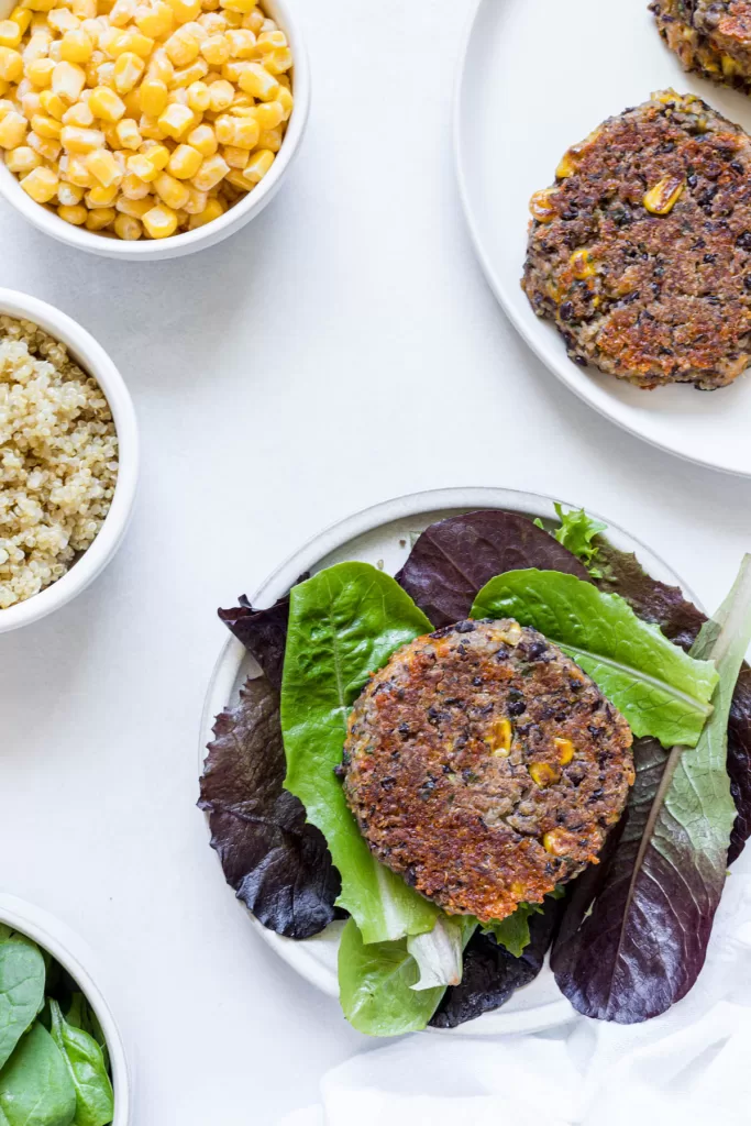 An overhead view of a black bean fritter on a bed of lettuce with a bowl of quinoa and corn next to it.