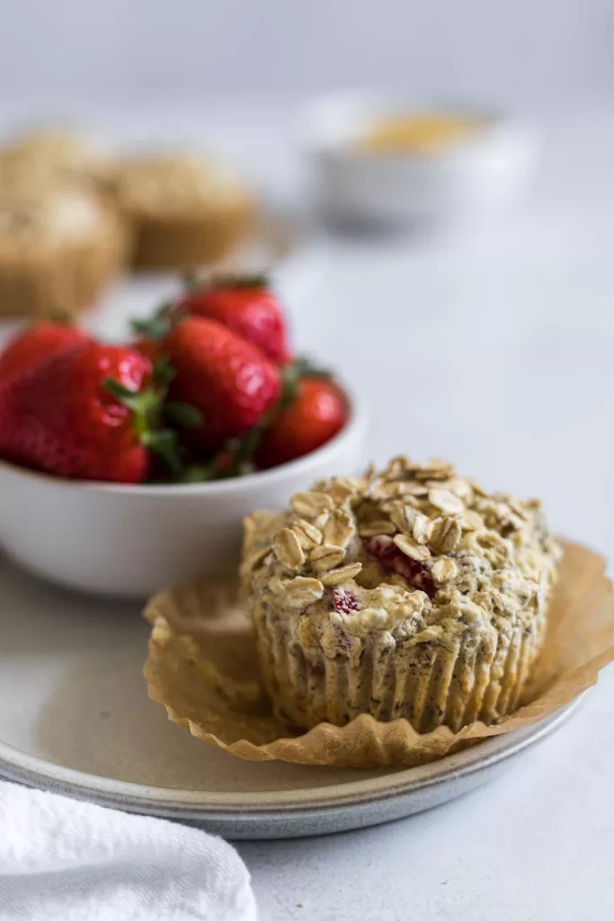 A Gluten-Free Strawberry Muffin on a plate with a bowl of strawberries behind it.