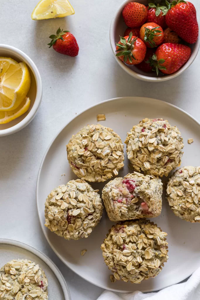 A group of strawberry muffins on a plate with a bowl of strawberries and lemons next to it.