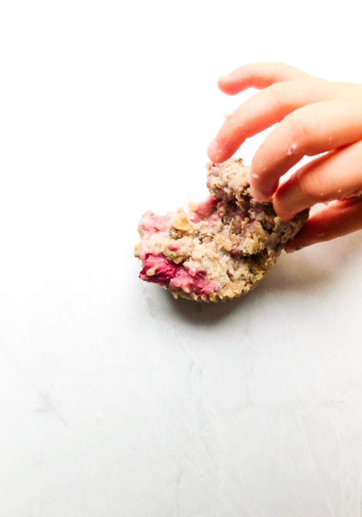 A toddler's hand is reaching for a half eating strawberry muffin.