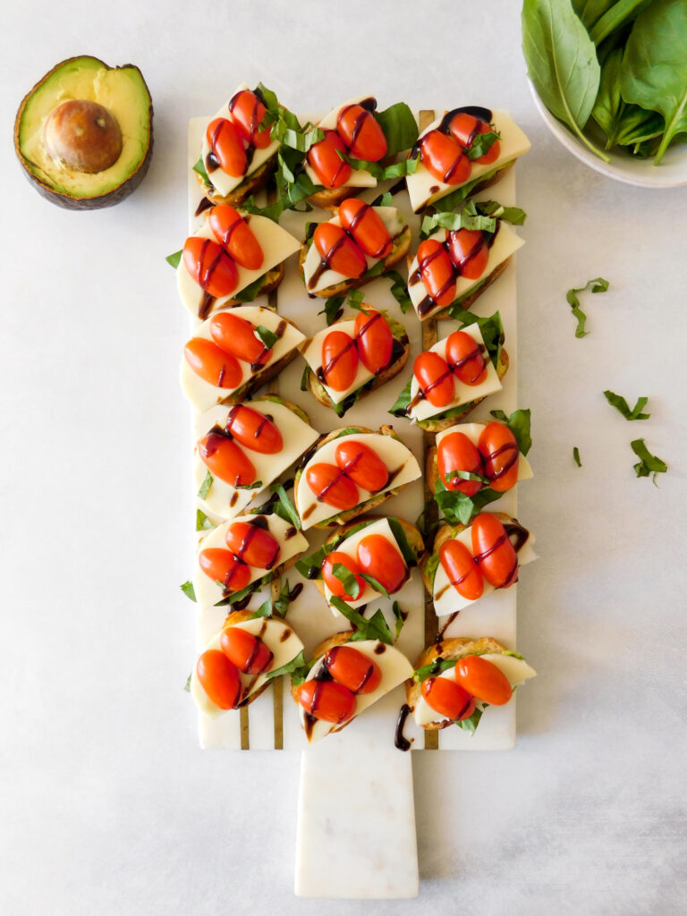 Caprese Crostini sit in rows on a white marble serving board. An open avocado and basil leaves sit to the side of the board.