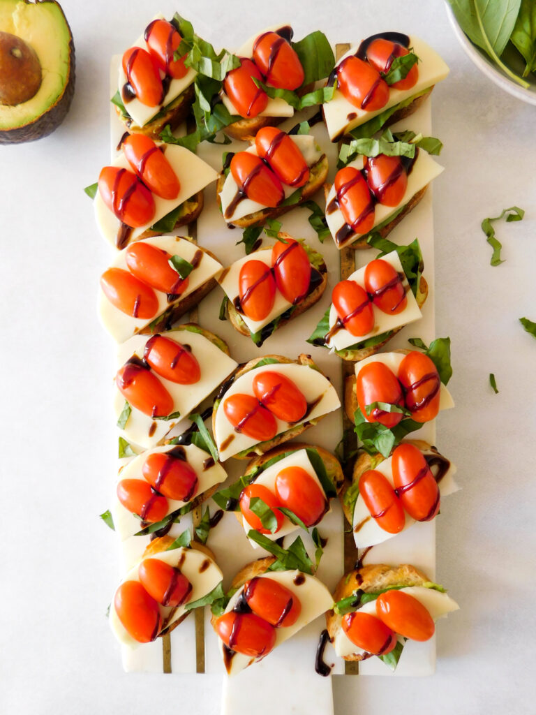 Crostini sit in rows on a white marble serving board. An open avocado and basil leaves sit to the side of the board.