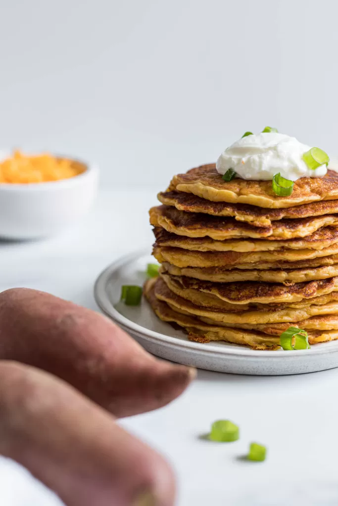A stack of pancakes with sweet potatoes next to it and a bowl of shredded cheese behind it.