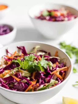 Dairy-Free Coleslaw in two bowls. Some lemon wedges and cilantro sit next to it. Two small bowls of cabbage and carrots sit in the background.