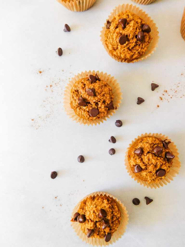 Four oat flour pumpkin muffins are arranged vertically with chocolate chips scattered around them.
