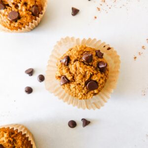 A Healthy Oat Flour Pumpkin Muffin with chocolate chips on top of it sits on top of a muffin liner that is frayed out. Two other muffins sit near by and chocolate chips are scattered around.