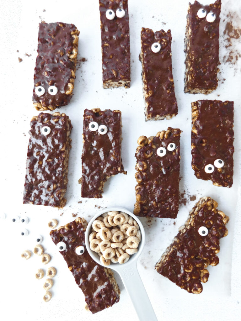 Monster cheerio bars are arranged flat and spread out from each other. One of them has a big bite taken out of it. A cup of cheerios is in the middle of the image along with some cheerios and candied eyeballs strewn within the cereal bars.