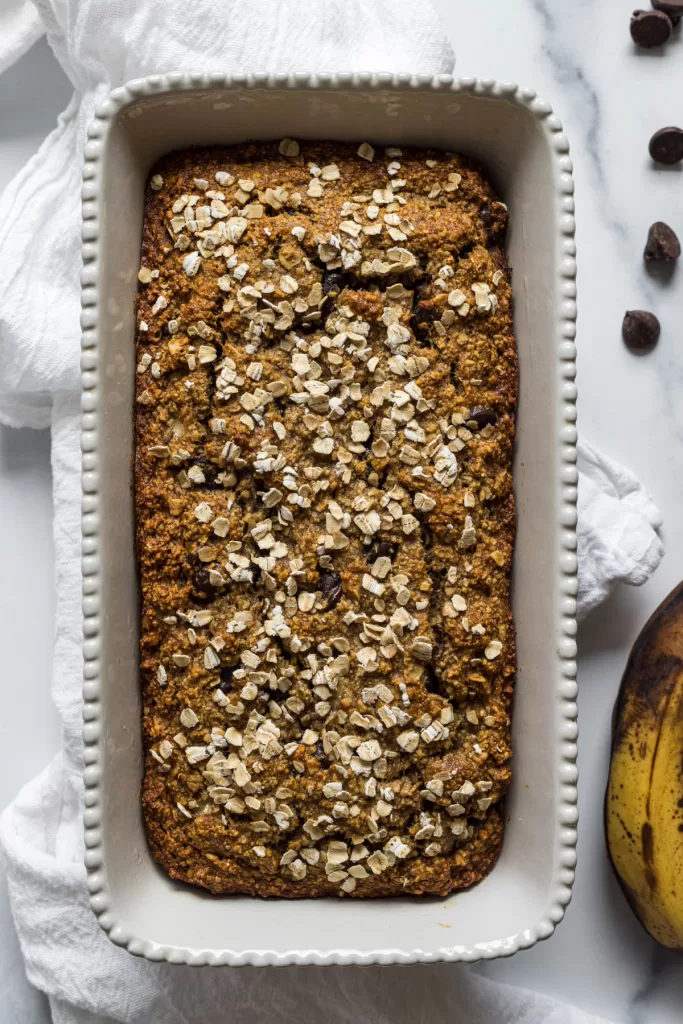 A loaf of banana bread on a white cloth with a ripe banana and chocolate chips next to it.