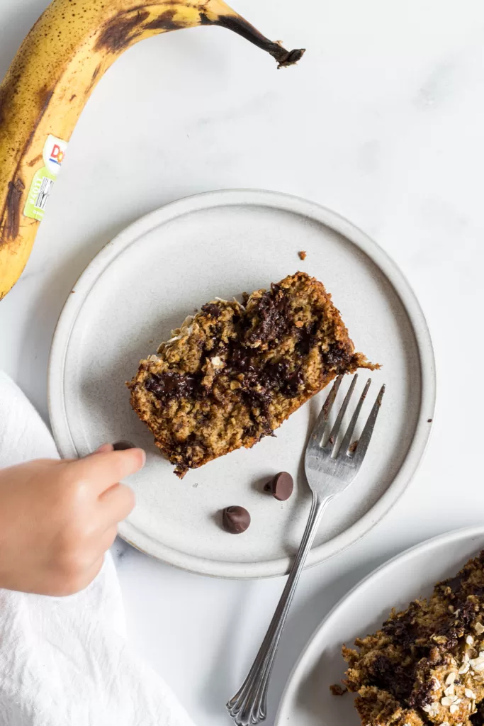 A toddler's hand is reaching for a chocolate chip on a plate where there is a slice of banana bread.