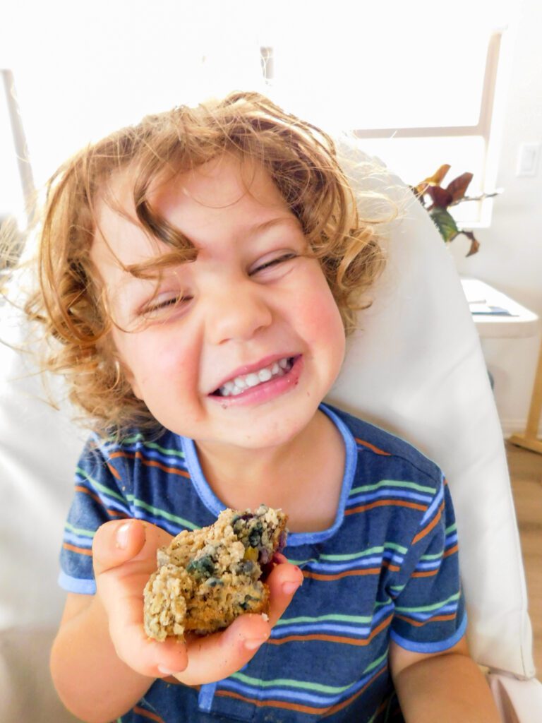 A two-year-old boy is sitting in a high chair holding a half eaten muffin and smiling at the camera.