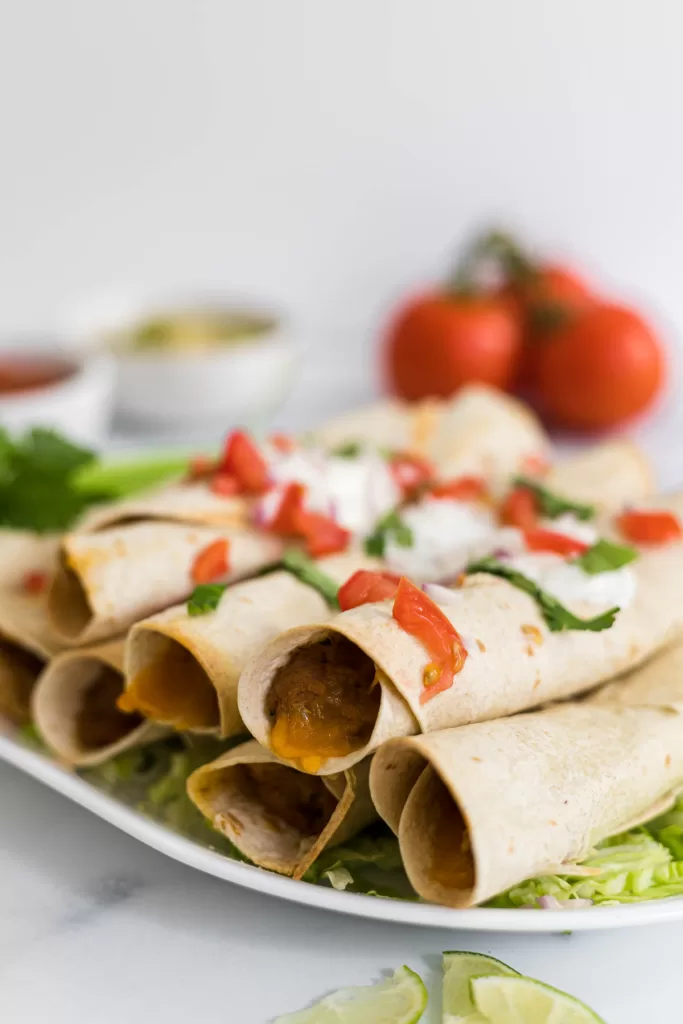 A side view of baked flautas on a plate with sour cream and tomatoes on top.