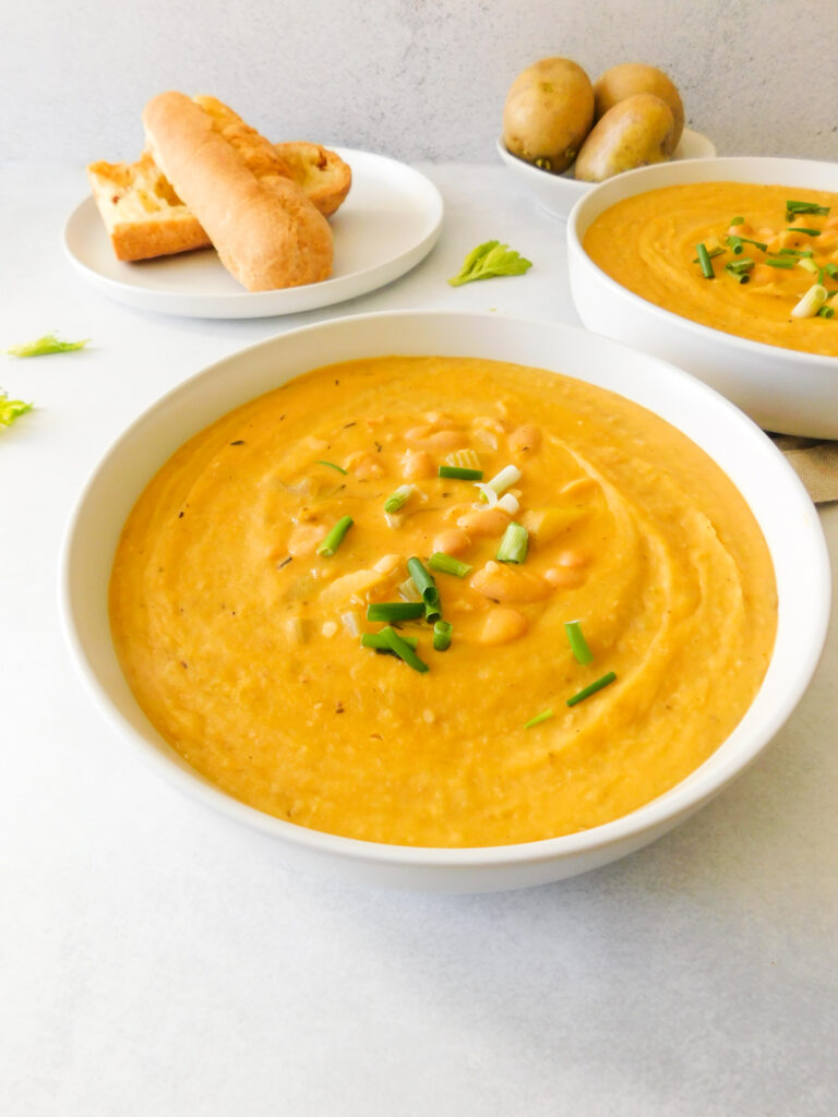 A side view of two bowls of White Bean Potato Soup. A plate with a baguette sits in the background and a bowl with potatoes.