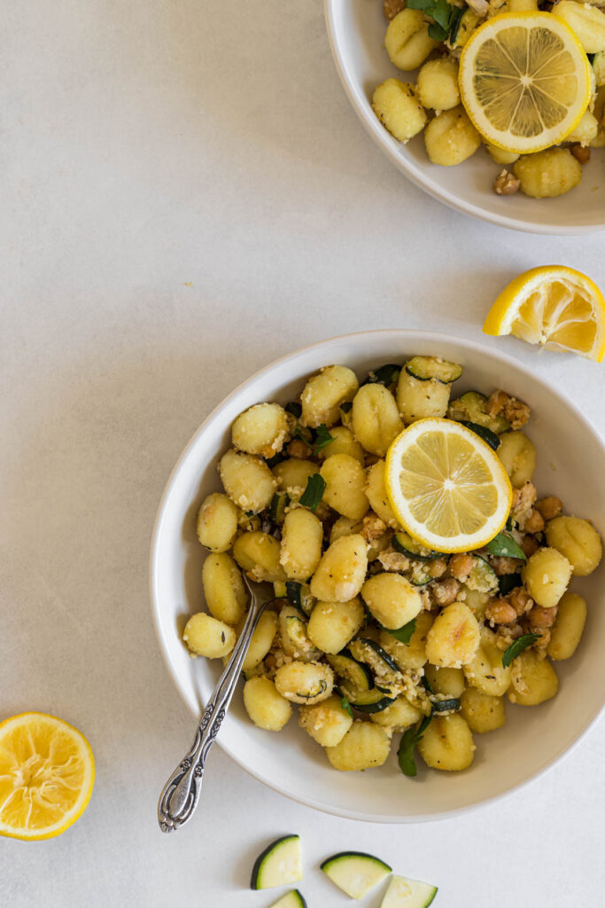 A bowl of gnocchi. Another bowl peaks into the top of the frame. Some lemon wedges and zucchini slices sit next to the main bowl.