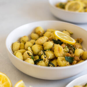 A bowl of Sheet Pan Gnocchi With Roasted Zucchini and Chickpeas. Two more bowls sit next to it and some lemon wedges are off to the side.