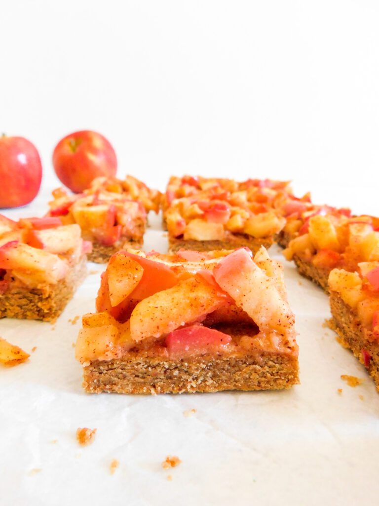 A close up view of the side of an apple pie bar. More apple pie bars sit behind it as well as two apples.