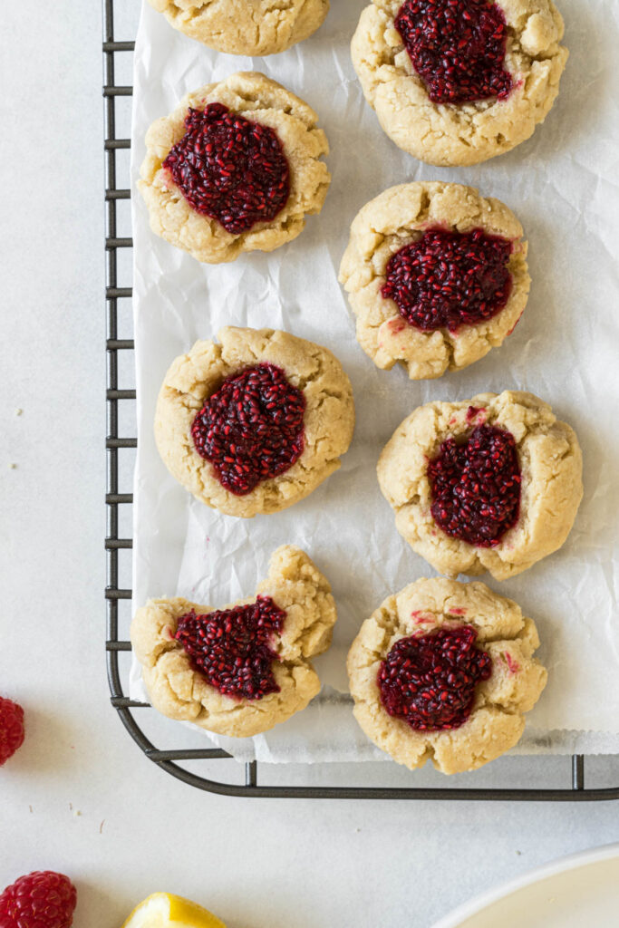An overhead view of raspberry thumbprint cookies sitting on a wire rack. Some raspberries, a lemon wedge, and a plate sit to the side.