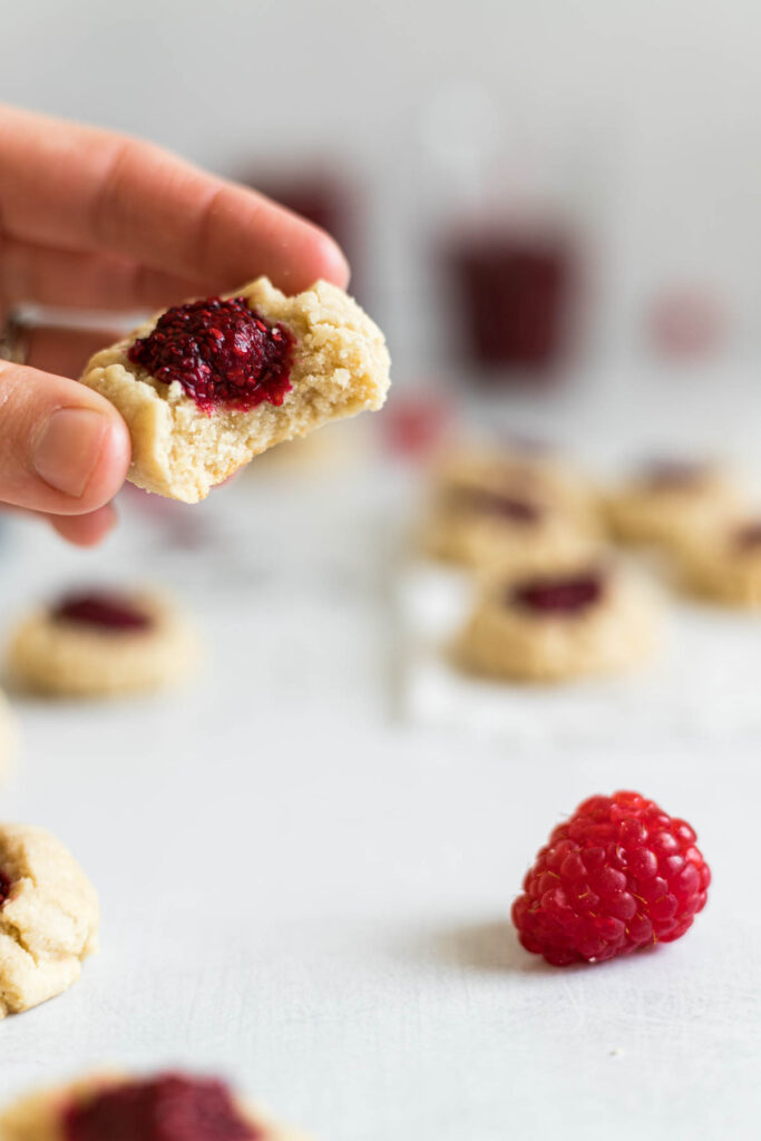A white hand is picking up a raspberry cookie with a bite out of it. Below it sits a raspberry. In the background there are more cookies and two jam jars.