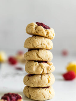 A stack of six Vegan Thumbprint Cookies With Raspberry Chia Jam. A cookie with a bite out of it sits in the front of the stack. Behind it are some raspberries and a lemon wedge.