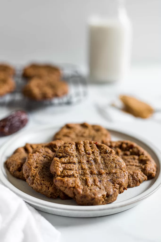 A plate of peanut butter date cookies. Some more cookies sit behind it, as well as a glass of milk, a spoonful of peanut butter, and dates.