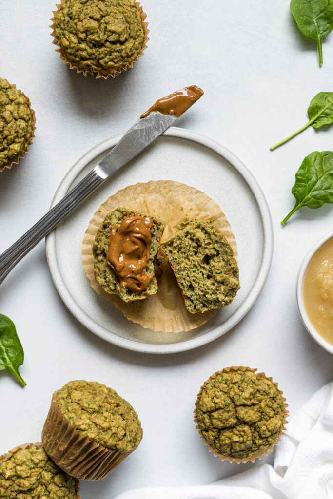A spinach banana muffin cut in half on a plate. One half has nut butter smeared on it and a knife sits next to it.