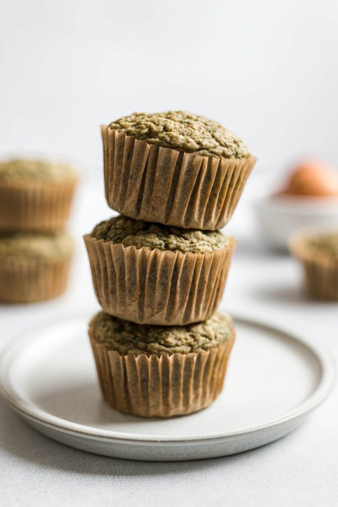 Three Banana Spinach Muffins are stacked on top of a small plate. More muffins and a bowl with eggs sit in the background.
