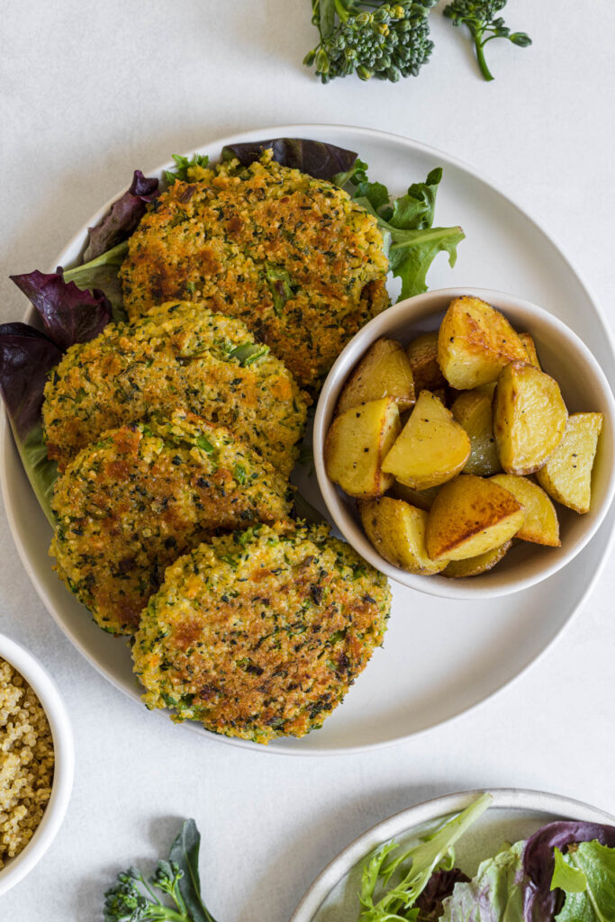 An overhead view of a plate of Broccoli Quinoa Patties on a bed of lettuce. A small bowl of roasted potatoes sit next to it. A salad plate is peaking into the image as well as a small bowl of quinoa.