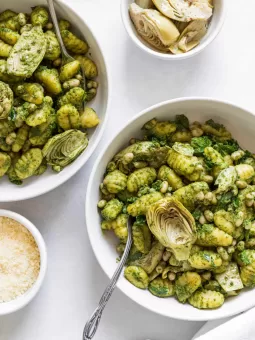 Two bowls of Sheet-Pan Pesto Gnocchi with a bowl of artichokes and parmesan next to it.