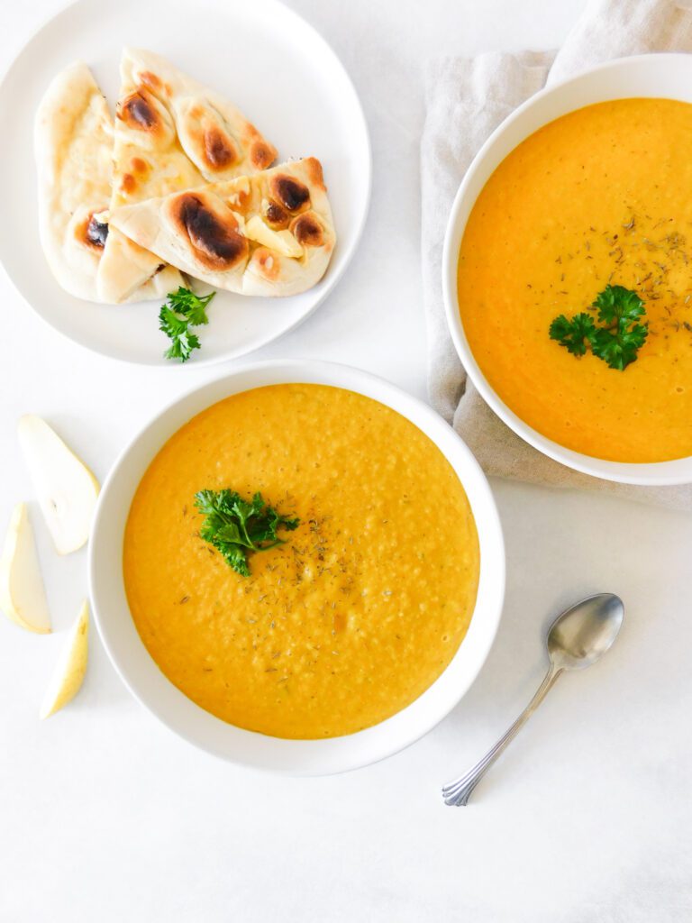 An overhead view of two bowls of Curried Butternut Squash and Pear Soup. A plate with naan bread sits in the top left of the image and slices of pear sit next to the soup. 