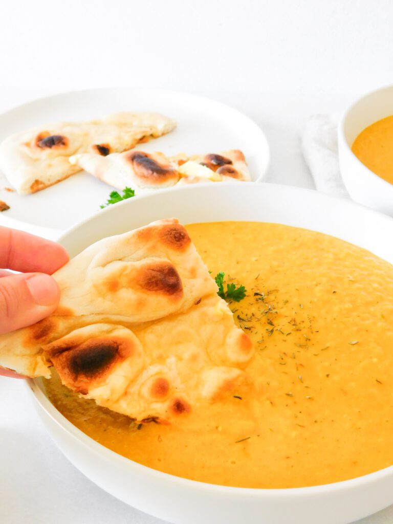 Naan bread is being dipped into a bowl of Curried Butternut Squash and Pear Soup. More naan bread and soup sit in the background.