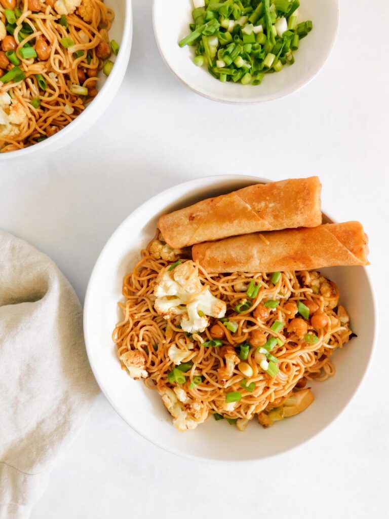 A bowl of Easy Vegan Ramen Noodles with spring rolls on the side of the bowl. Part of another bowl of ramen and some green onions are shown as well.