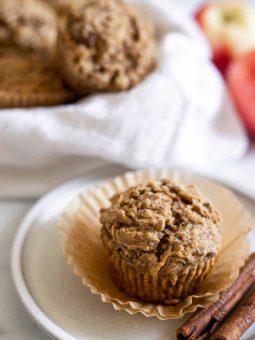 A Vegan Apple Cinnamon Muffin on a plate with cinnamon sticks next to it. A bowl of muffins is behind it as well as two apples.