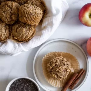 An overhead view of the muffins with one on a plate and more in a bowl. A bowl of chia seeds and some apples sit next to the plate.