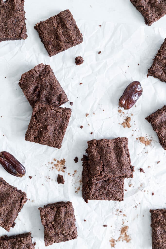 Vegan Date Brownies are scattered throughout the image. Some are stacked on one another. There is cacao powder and dates sprinkled throughout.