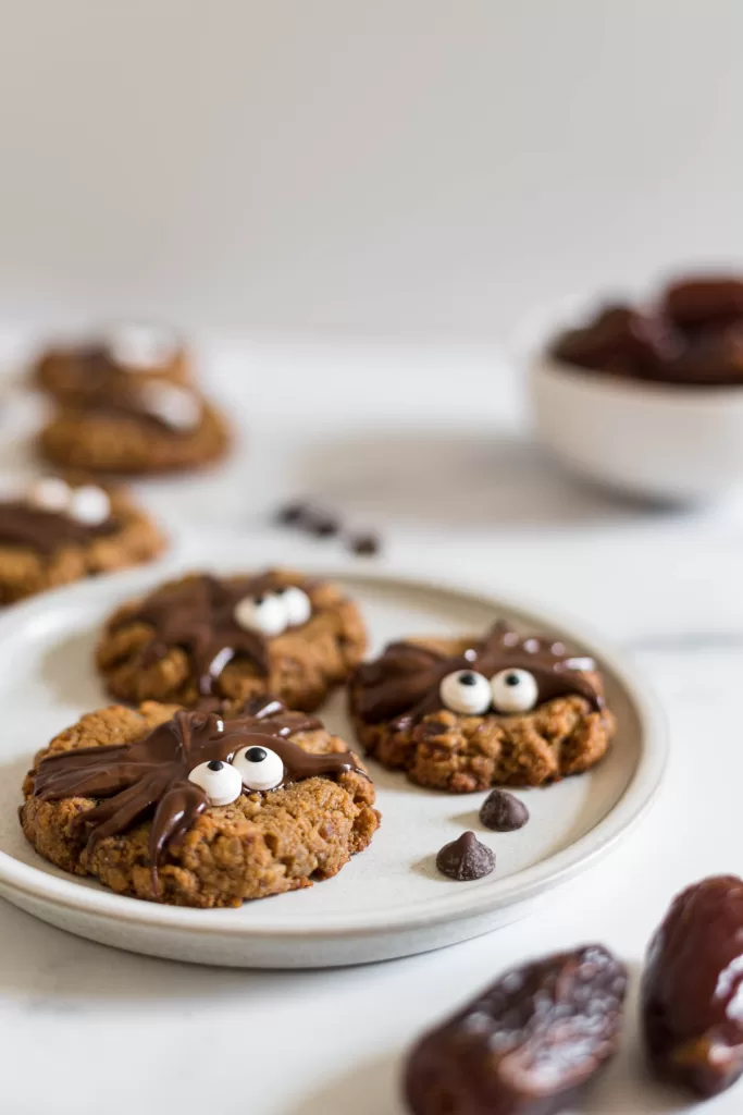 Three peanut butter date spider cookies on a plate with a bowl of dates and more cookies behind it.