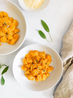 Two bowls of Sheet-Pan Gnocchi with Pumpkin Pasta with several sage leaves surrounding them. A tan colored linen cloth sits on the right side of the bowl that's sitting in the center of the image.