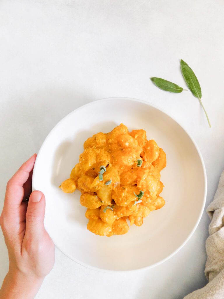 Kaileigh's left hand is resting on the side of a bowl of pumpkin gnocchi. Two sage leaves and a glimpse of a tan linen cloth sit to the right side of the bowl. 