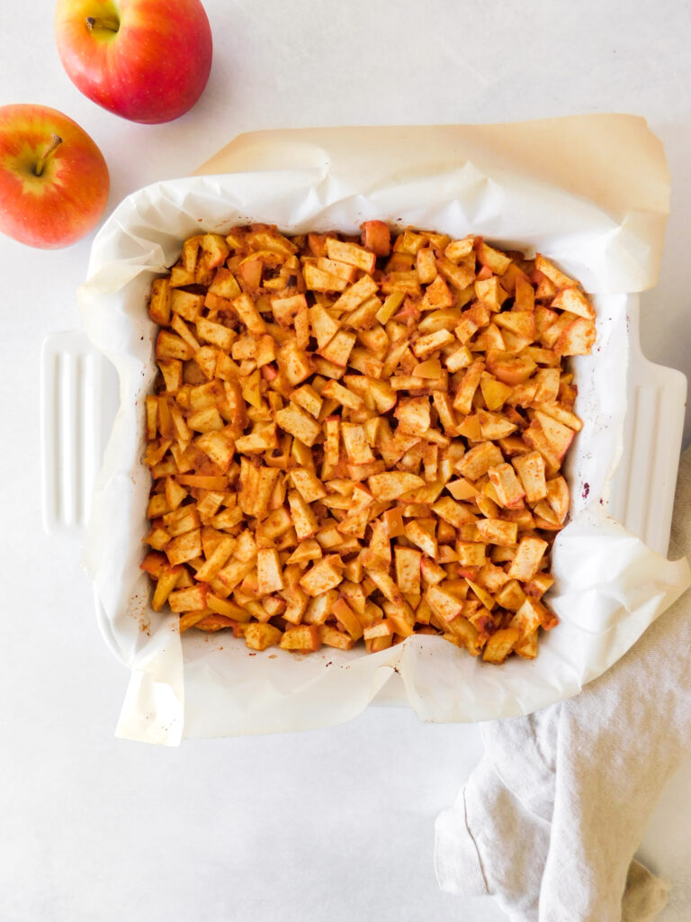 Apple Pie Bars are in a baking dish, uncut, with two apples sitting next to it.