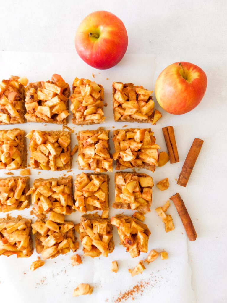 Apple pie bars sit in rows on parchment paper with two apples and cinnamon sticks surrounding it.