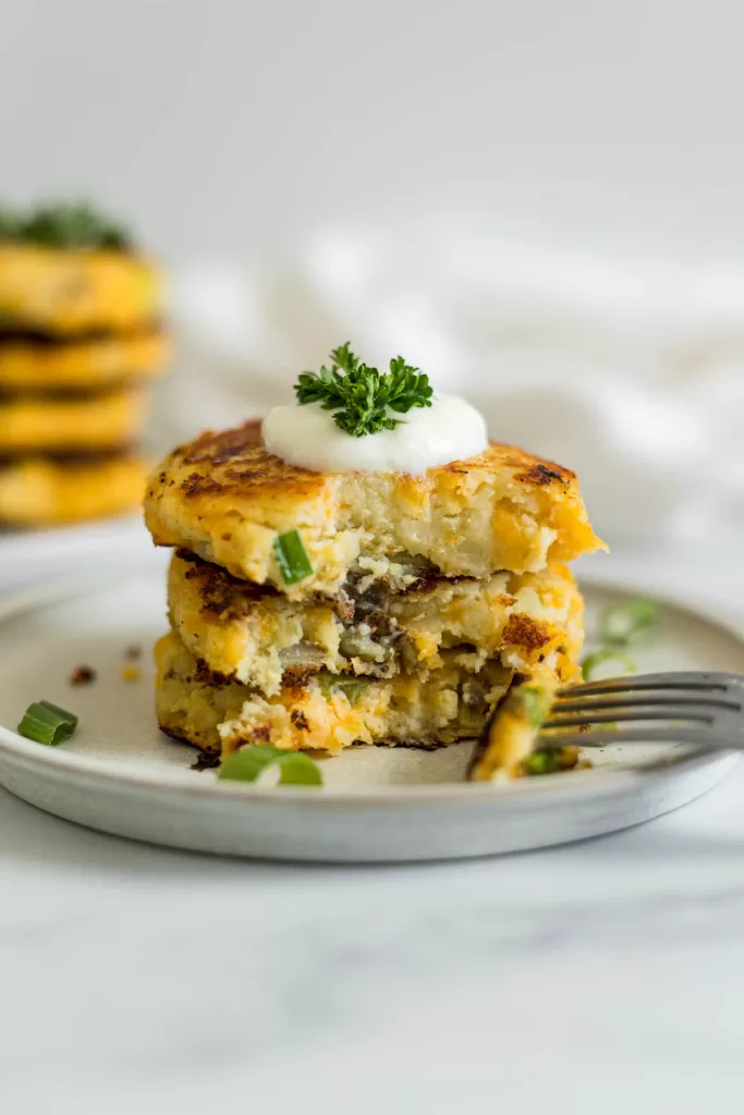A stack of three mashed potato pancakes. A fork has a piece of the pancakes on it and is lying next to the pancake stack on a plate.
