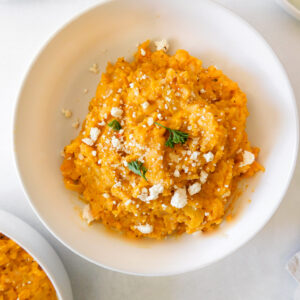A bowl of roasted butternut squash risotto with feta crumbles sprinkled on top and some parsley leaves.